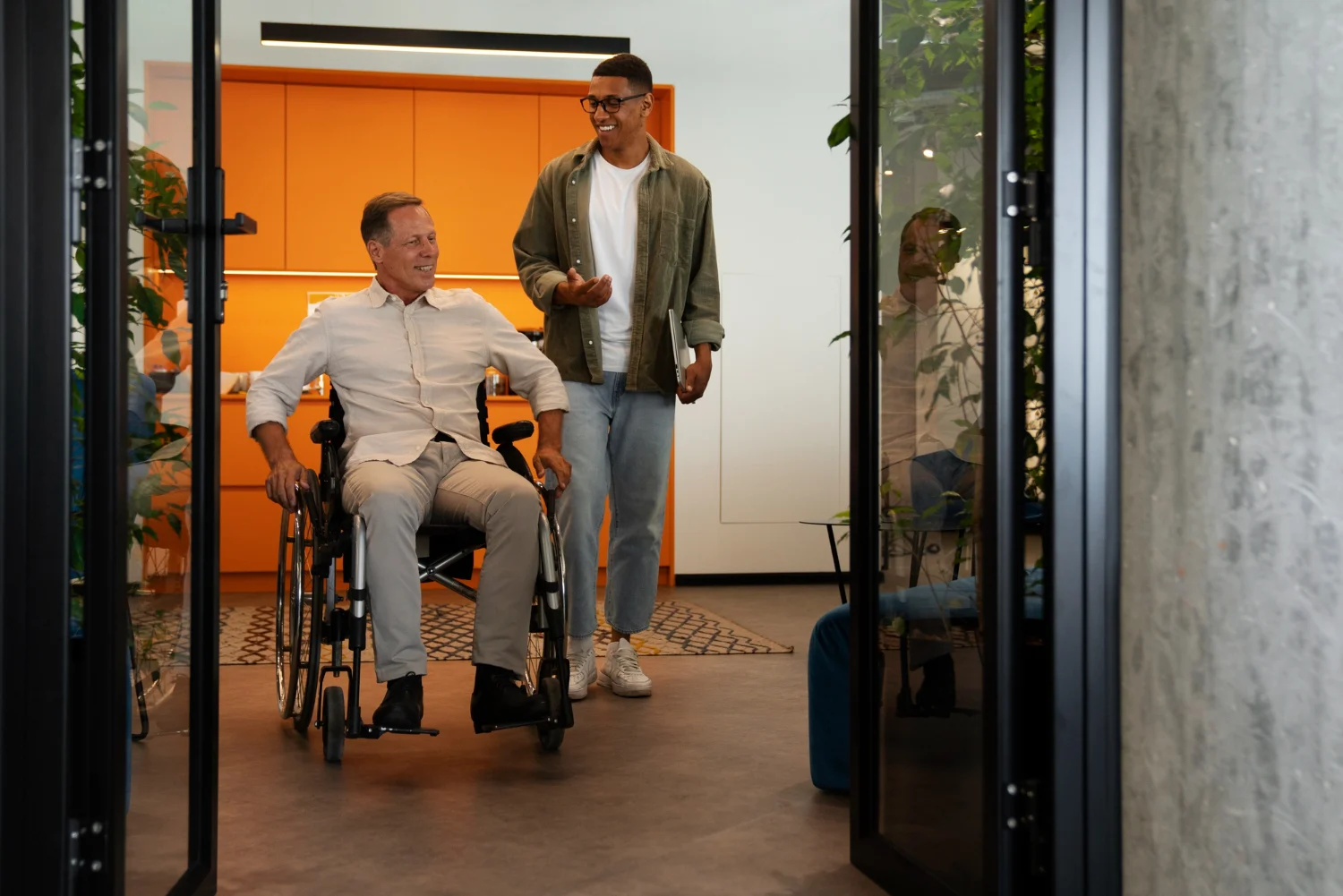 Man in a wheelchair and a colleague passing through a wide glass door opening in an office
