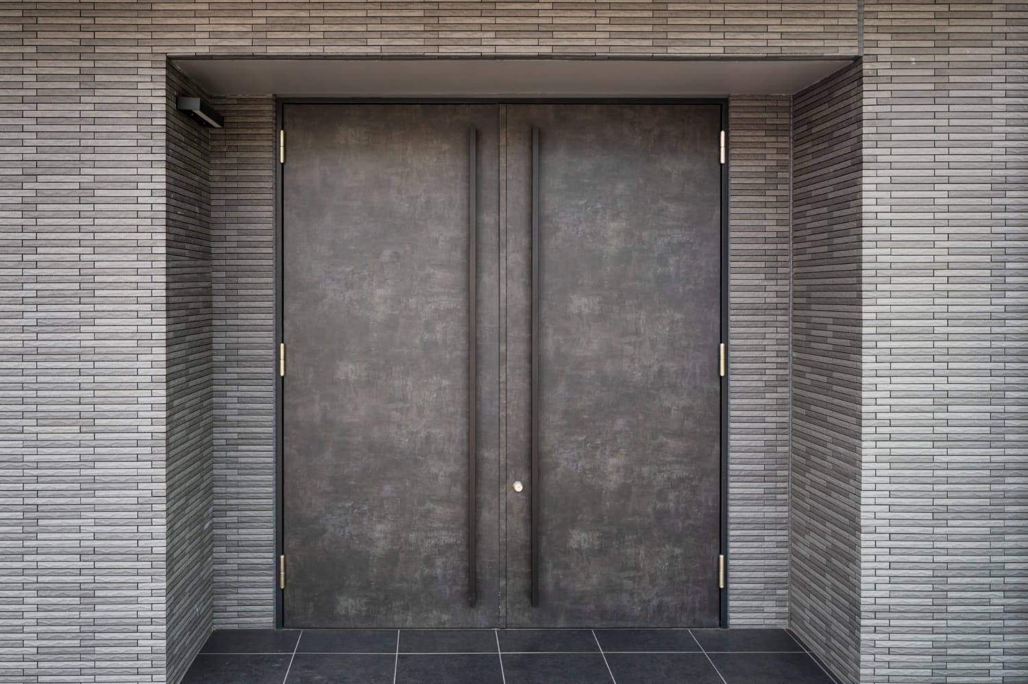 Dark textured double entry doors with long vertical pull handles set in a gray brick wall
