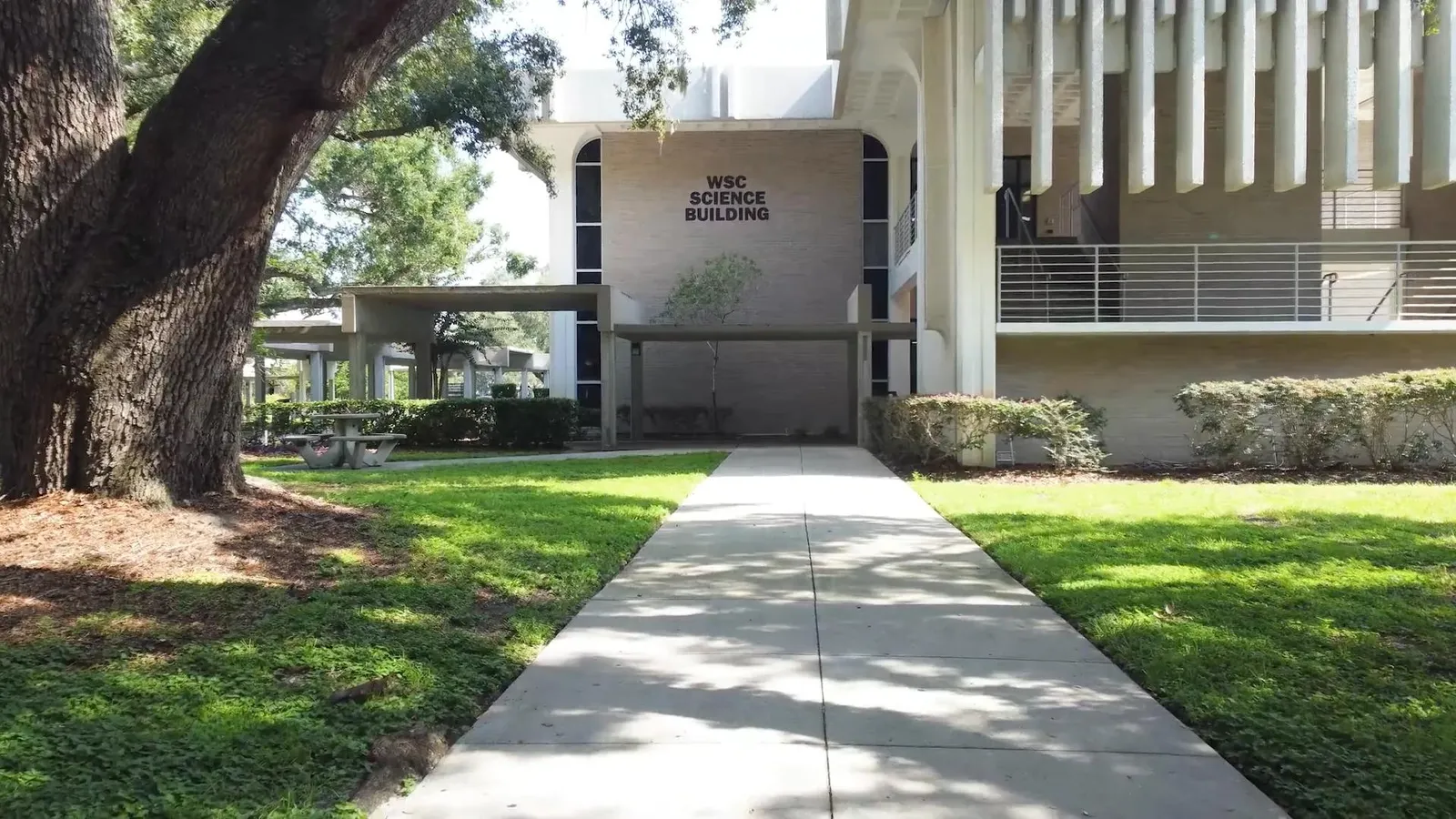 Entrance walkway to the WSC Science Building at Polk State College in Winter Haven, Florida, with the building name on a stucco wall and a large oak tree